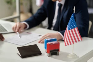 Immigration officer reviewing paperwork at a desk with a U.S. flag, passport, and ‘approved’ and ‘rejected’ stamps.